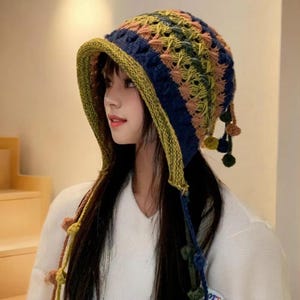May include: A multi-colored crochet hat featuring a wide brim and dangling pom-poms. The hat is constructed with a textured pattern in shades of navy blue, olive green, and soft peach, finished with a solid olive green border along the brim edge.