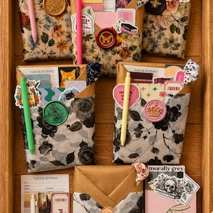 Puede incluir: Cinco sobres de papel rectangulares con patrones florales, cada uno sellado con cera y que contiene tarjetas de rese&ntilde;as de libros, pegatinas y bol&iacute;grafos. Incluye un bol&iacute;grafo blanco, un marcap&aacute;ginas Book Festival y un cuaderno rosa con cerezas.