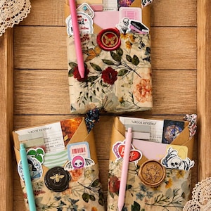 May include: Three rectangular paper-wrapped packages featuring floral patterns in shades of red, orange, and green. Each package is sealed with a wax stamp, decorated with various stickers, and includes a book review card and a colored ballpoint pen.