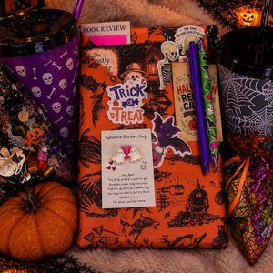 May include: A Halloween-themed gift set featuring an orange book wrapped in bat-patterned paper, a purple skull-print tumbler, a black spiderweb-print tumbler, a small pumpkin, two pens, a Ghostie Pocket Hug card, and various spooky stickers.