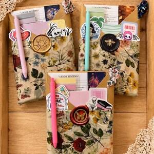 Puede incluir: Tres sobres rectangulares envueltos en papel floral, cada uno con una tarjeta de rese&ntilde;a de libro, pegatinas y un bol&iacute;grafo. Los sobres est&aacute;n sellados con lacre con motivos de sol, abeja y mariposa, e incluyen peque&ntilde;as gomas de tela.