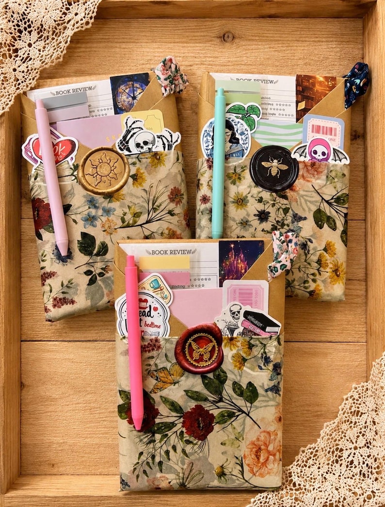 Puede incluir: Tres sobres rectangulares envueltos en papel floral, cada uno con una tarjeta de rese&ntilde;a de libro, pegatinas y un bol&iacute;grafo. Los sobres est&aacute;n sellados con lacre con motivos de sol, abeja y mariposa, e incluyen peque&ntilde;as gomas de tela.