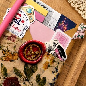 Puede incluir: Un sobre con estampado floral sellado con un sello de cera rojo que muestra una mariposa dorada. Dentro hay pegatinas, un bol&iacute;grafo rosa, una nota adhesiva amarilla de rese&ntilde;a de libro y un ticket. El sobre muestra ilustraciones bot&aacute;nicas.