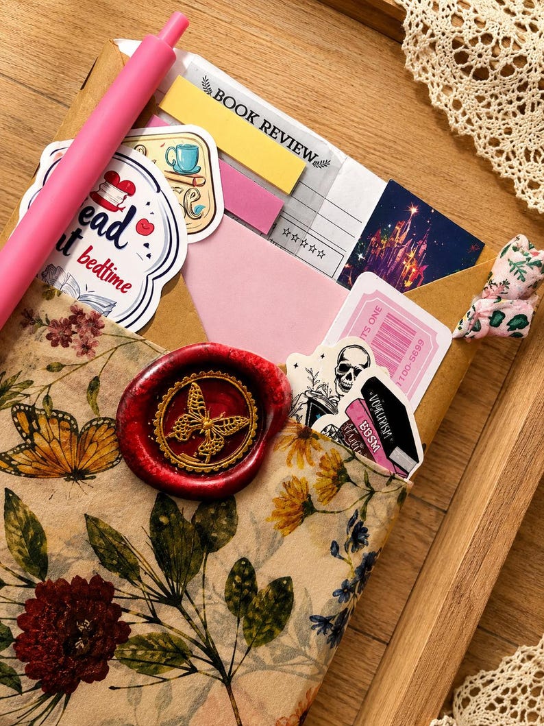 Puede incluir: Un sobre con estampado floral sellado con un sello de cera rojo que muestra una mariposa dorada. Dentro hay pegatinas, un bol&iacute;grafo rosa, una nota adhesiva amarilla de rese&ntilde;a de libro y un ticket. El sobre muestra ilustraciones bot&aacute;nicas.