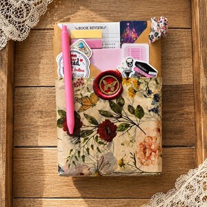 Puede incluir: Un paquete rectangular envuelto en papel floral con mariposas y flores, sellado con un sello de cera roja con forma de mariposa. Incluye un bol&iacute;grafo rosa brillante, una tarjeta de rese&ntilde;a de libros y pegatinas de una calavera y libros.