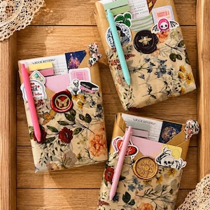 Puede incluir: Tres paquetes rectangulares envueltos en papel floral, cada uno con una tarjeta de rese&ntilde;a de libro, pegatinas y un bol&iacute;grafo. Cada paquete est&aacute; sellado con un sello de cera con dise&ntilde;o de mariposa, abeja o sol y un peque&ntilde;o lazo de tela.