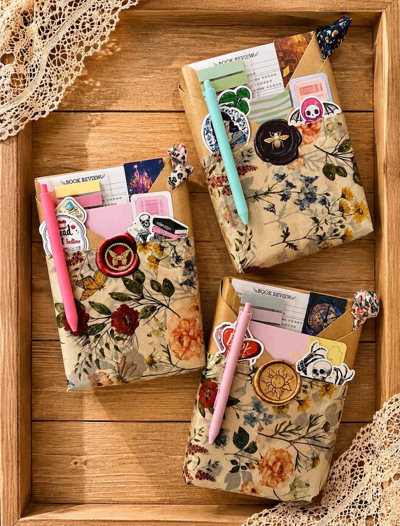 Puede incluir: Tres paquetes rectangulares envueltos en papel floral, cada uno con una tarjeta de rese&ntilde;a de libro, pegatinas y un bol&iacute;grafo. Cada paquete est&aacute; sellado con un sello de cera con dise&ntilde;o de mariposa, abeja o sol y un peque&ntilde;o lazo de tela.