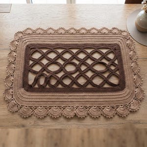 May include: A rectangular crochet placemat featuring a dark brown center with an openwork lattice pattern, surrounded by concentric borders in light tan and beige. The outer edge is finished with a decorative scalloped crochet border.