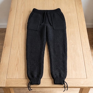 May include: A pair of black crochet joggers featuring a ribbed elastic waistband with a drawstring, two front pockets, and ribbed ankle cuffs that include adjustable drawstring ties at the bottom of each leg for a secure and fitted appearance.