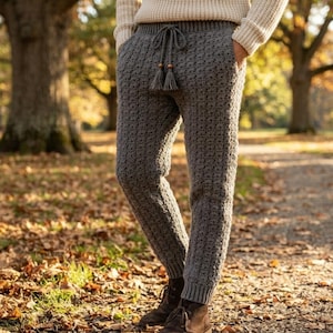 May include: A pair of charcoal gray crocheted pants featuring a textured stitch pattern, a ribbed waistband with a drawstring closure, and two decorative tassels. The pants are paired with a cream-colored long-sleeved knit sweater and brown boots.