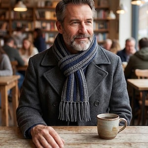 May include: A charcoal gray wool coat with a double-breasted front and a thick, ribbed knit scarf featuring alternating vertical stripes of gray and navy blue with long, knotted fringe ends, paired with a speckled beige ceramic mug.