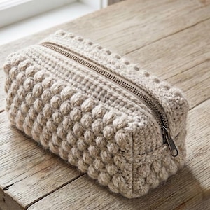 Puede incluir: Un estuche rectangular de ganchillo en color beige con un patrón de puntos de burbuja texturizados en los laterales y la parte superior. El estuche se cierra con una cremallera metálica a lo largo del borde superior, ofreciendo una apertura.