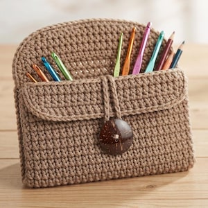 May include: A tan crochet organiser pouch featuring a rounded flap, a dark brown circular button closure, and a series of colourful metal crochet hooks and pencils inserted into the top compartment, showcasing a textured, hand-stitched yarn construction.