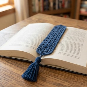 Peut inclure: Un marque-page au crochet bleu foncé avec un motif ajouré et un pompon en fil assorti à l'extrémité, posé dans un livre ouvert à couverture rigide aux pages blanches avec du texte imprimé noir, présentant une forme rectangulaire et texturée.