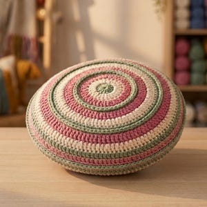 May include: A round, crocheted pillow featuring concentric circles in alternating shades of dusty rose, sage green, and cream. The textured yarn surface displays a consistent stitch pattern radiating from the center to the outer edge of the cushion.