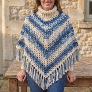 May include: A crochet poncho featuring a high turtleneck collar and a V-shaped hemline. The garment displays a repeating pattern of cream and variegated blue yarn in a granny square stitch, finished with long cream fringe along the bottom edge.