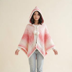 May include: A hooded crochet poncho featuring an ombre colour gradient transitioning from white to soft pink and deep rose. The garment has an open front with a drawstring closure finished with two white pom-poms, a textured stitch pattern, and a scalloped edge.