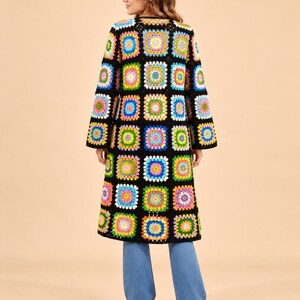 Könnte beinhalten: Ein langer Cardigan mit einem lebendigen Granny-Square-Häkelmuster in verschiedenen Farben wie Gelb, Blau, Pink und Grün, die mit schwarzem Garn verbunden sind. Das Kleidungsstück hat lange Ärmel und eine gerade, vorne offene Silhouette.