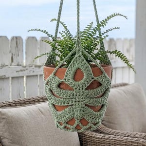 May include: A sage green crocheted plant hanger featuring a monstera leaf pattern that encases a terracotta pot containing a green fern. The hanger includes long crocheted cords for suspension, creating a decorative holder for the plant.