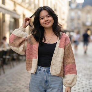 May include: A color-blocked crochet cardigan featuring horizontal panels in shades of cream, tan, and dusty rose. The garment has long, voluminous sleeves with ribbed cuffs, a V-neckline, and a button-down front, worn over a black top and blue jeans.