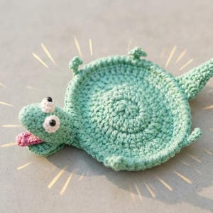 May include: A light green crocheted coaster shaped like a turtle with a circular shell body, a small head with white and black eyes, a pink tongue sticking out, four short legs, and a pointed tail, accented by radiating graphic lines.