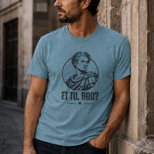 May include: A heathered blue short-sleeve t-shirt featuring a black graphic print of a Roman figure wearing a laurel wreath inside a circle. Below the graphic, the text reads ET TU, BRO? with a small star centered between two horizontal lines.