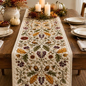 May include: A quilted table runner featuring a repeating pattern of autumn leaves, acorns, and clusters of red berries on a cream-colored background. The runner has a diamond-stitched texture and is centered on a wooden dining table with place settings.