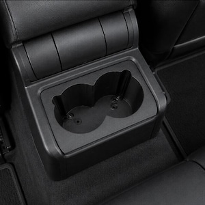 May include: A black plastic dual cup holder console designed for a vehicle interior. The unit features two circular recessed compartments with visible mounting screws at the base, integrated into a rectangular housing with a smooth matte finish.