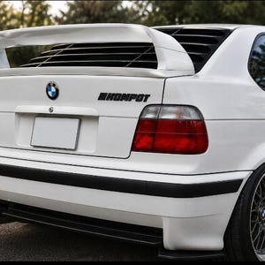 May include: A white BMW hatchback featuring a large rear spoiler, black window louvers, and a black decal reading KOMPOT on the trunk lid. The vehicle has red and clear taillights, a black bumper trim, and dark multi-spoke wheels with silver rims.