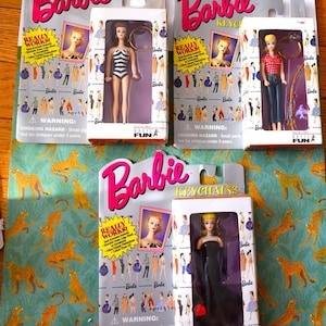 May include: Three Barbie keychain dolls in original packaging. One doll wears a black and white striped swimsuit, one wears a red and white checkered shirt with blue jeans, and one wears a black evening gown. Each box features the Barbie logo.