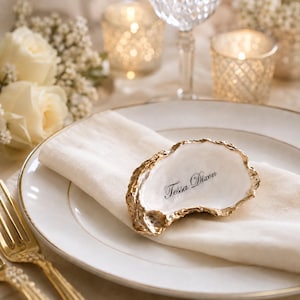 May include: A white oyster shell place card with gold-painted edges, featuring the name Tessa Dixon printed in black script. The shell rests on a folded cream-coloured fabric napkin atop a white dinner plate with a thin gold rim.
