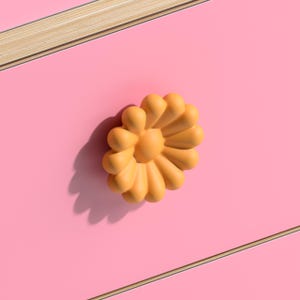 Op de afbeelding: Een feloranje, bloemvormige ladeknop gemonteerd op een glad, effen roze oppervlak. De knop heeft afgeronde, bloembladachtige segmenten die vanuit een centraal punt uitstralen en een duidelijke schaduw werpen op het roze paneel.