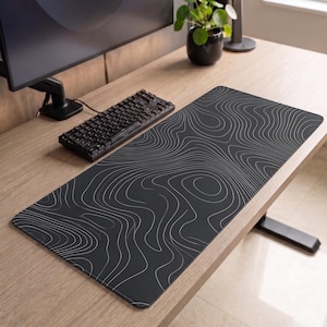 Peut inclure: Un grand tapis de bureau rectangulaire doté d'une surface noire avec un motif de lignes topographiques blanches. Le tapis présente des coins arrondis et une finition lisse, offrant une large zone pour les périphériques informatiques.