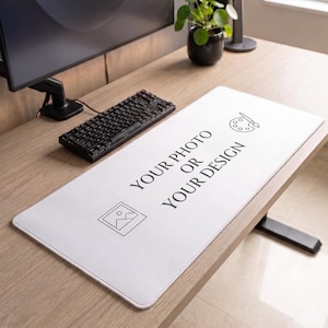 Op de afbeelding: Een rechthoekige witte bureaumat met een zwart lijntekening-icoon van een fotolijst, een verfpalet met een penseel en de gedrukte tekst "YOUR PHOTO OR YOUR DESIGN" in een gecentreerd, schreefloos lettertype op het gladde oppervlak.