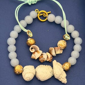 Può includere: Un bracciale con perline azzurre, sfere dorate testurizzate e tre grandi conchiglie bianche. Un secondo filo più piccolo include perline azzurre, sfere dorate e piccole conchiglie marroni e bianche con una chiusura a T dorata.