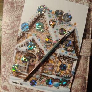 Op de afbeelding: Een rechthoekige kaart met een illustratie van een peperkoekhuisje, versierd met talloze ronde, iriserende, holografische stickers en de tekst Totaal 40. Een dunne, metalen, donkergekleurde stylus ligt diagonaal over het oppervlak van de kaart.