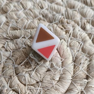 Op de afbeelding: Een witte vierkante ring met twee driehoekige inlays, één in een effen bruine kleur en de andere in een effen koraalroze kleur. De ring heeft een gladde, matte afwerking en is in een hoek geplaatst om het geometrische ontwerp te tonen.