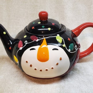 May include: A ceramic teapot with a snowman design. The teapot is black with a white face, orange nose, and a red handle and lid knob. It is decorated with colorful Christmas lights.