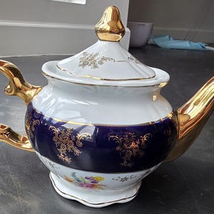 May include: A white porcelain teapot featuring a deep blue band around the center, decorated with gold floral filigree patterns. The teapot includes a gold-finished handle, spout, and lid finial, with additional small floral motifs near the base.
