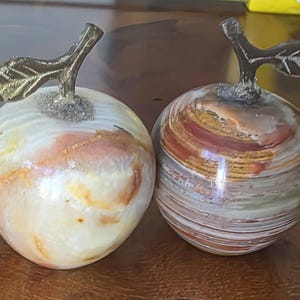 May include: Two decorative apple-shaped sculptures crafted from polished stone. The left apple features creamy white and orange swirls, while the right apple displays horizontal bands of brown, red, and white. Both have textured metal stems and leaves.