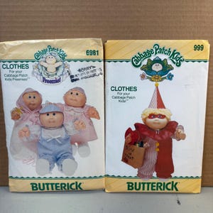 May include: Two Butterick sewing pattern envelopes for Cabbage Patch Kids clothes. The left envelope, number 6981, shows three dolls in baby outfits. The right envelope, number 999, features a doll in a red and white clown costume with a trick-or-treat bag.