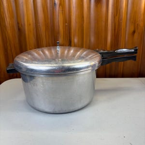 Puede incluir: Una olla a presión de aluminio de color plateado con una tapa circular que presenta una válvula metálica central. La olla tiene un mango largo de plástico negro que se extiende desde el lateral para un agarre seguro.