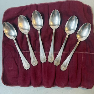 Puede incluir: Un juego de seis cucharas de metal plateado dispuestas en fila. Cada cuchara presenta un mango texturizado con un distintivo emblema en forma de escudo en la base. Las cucharas tienen cuencos ovalados lisos y superficies metálicas pulidas.