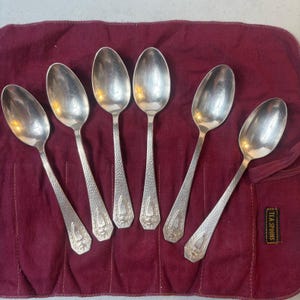 May include: Six silver-toned metal spoons with hammered handles and a decorative shield-shaped emblem at the base. The spoons are arranged on a dark red fabric roll with a small black tag that reads TEA SPOONS in gold lettering.