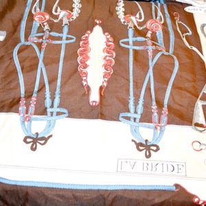 May include: A brown and cream silk scarf featuring a repeating equestrian print of light blue horse bridles, metal bits, and red accents. The text LA BRIDE is printed in the lower center, with additional French labels near various horse tack illustrations.