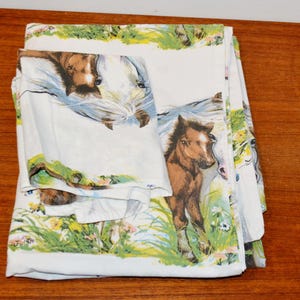 Op de afbeelding: Een wit stoffen laken met een herhalend patroon van bruine en witte paarden in groen gras met gele en witte bloemen. Het ontwerp bevat een horizontale rand van groen gebladerte en bloemen langs de boven- en onderkant van de stof.