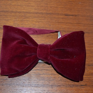 May include: A deep burgundy velvet bow tie featuring a soft, textured fabric surface and a central knot detail. The bow tie includes a matching adjustable fabric strap attached to the back, designed to secure the accessory around the neck.