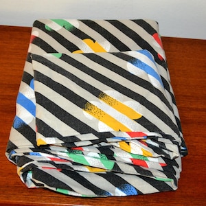 Pode incluir: Um lençol de tecido dobrado com um padrão de listras diagonais pretas e bege. O design é acentuado com formas florais estilizadas espalhadas em cores sólidas amarelo, azul, vermelho e verde, cada uma com um detalhe de textura pontilhada.