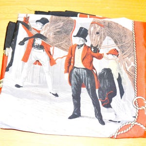 May include: A square silk scarf featuring a printed scene of figures in formal equestrian attire, including top hats and red coats, standing beside a brown horse. The design includes a rope border detail in white against a deep orange background.