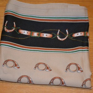 May include: A square scarf in a beige hue featuring a black horizontal band decorated with a repeating pattern of brown leather belts and white horseshoes. The lower section displays a repeating print of brown hunting horns on a beige background.